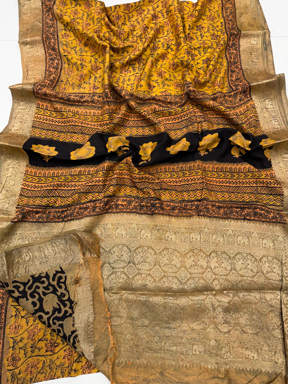 A traditional Indian saree laid flat, featuring a mustard yellow and black printed body with a wide beige and gold zari border, and a black border section decorated with gold floral motifs.