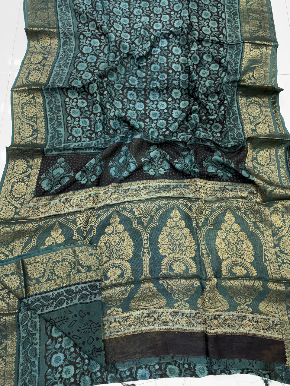 A dark teal and black Banarasi silk saree with intricate gold zari floral motifs and a wide decorative border, laid out on a white tiled floor.