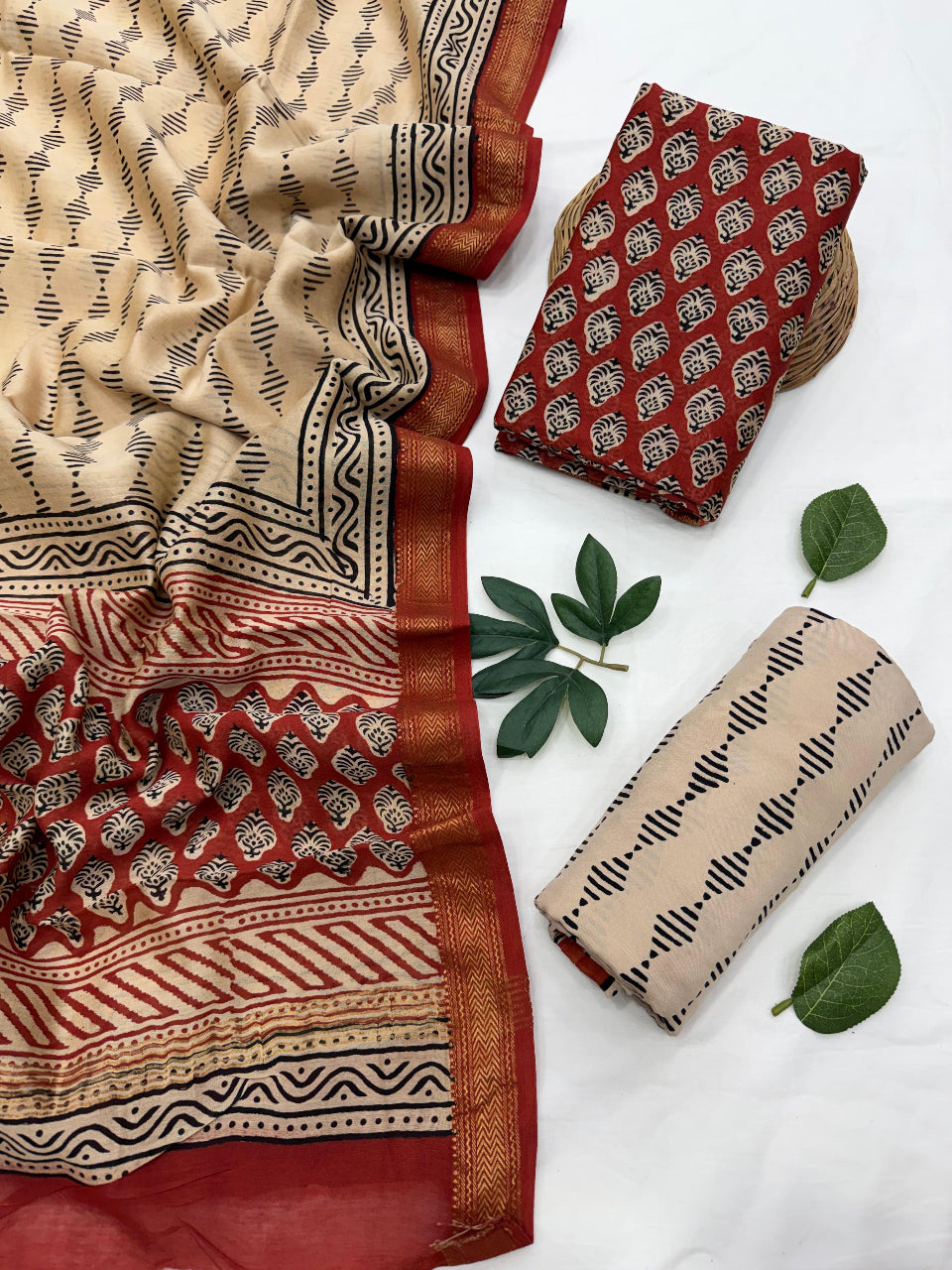 Beige and red patterned Maheshwari silk unstitched suit set with black geometric motifs and a red border, displayed with folded matching pieces and green decorative leaves on a white background