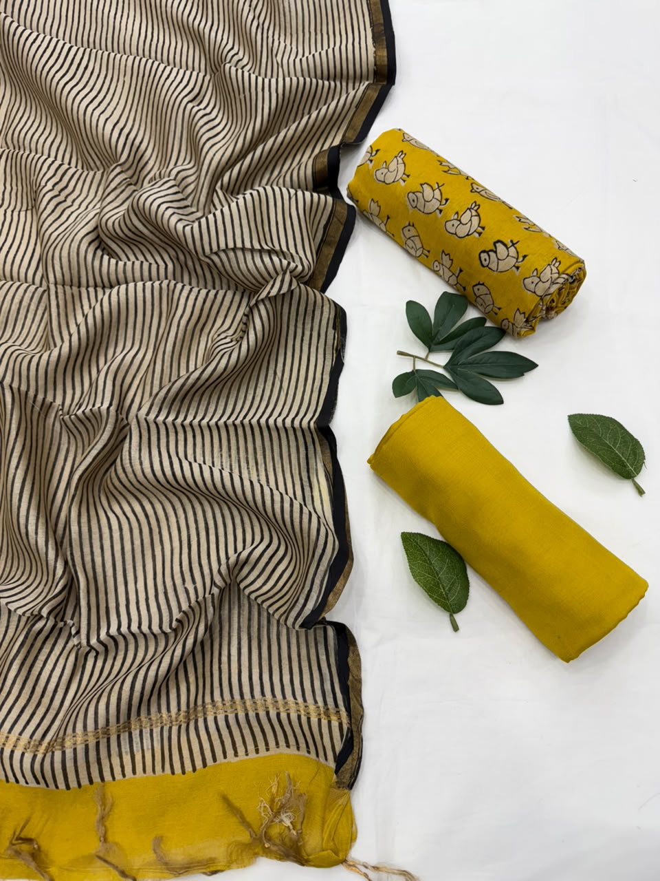 A folded and rolled mustard yellow fabric piece with a floral print and solid yellow sections, displayed alongside a matching striped beige and black fabric piece on a white background with decorative green leaves.