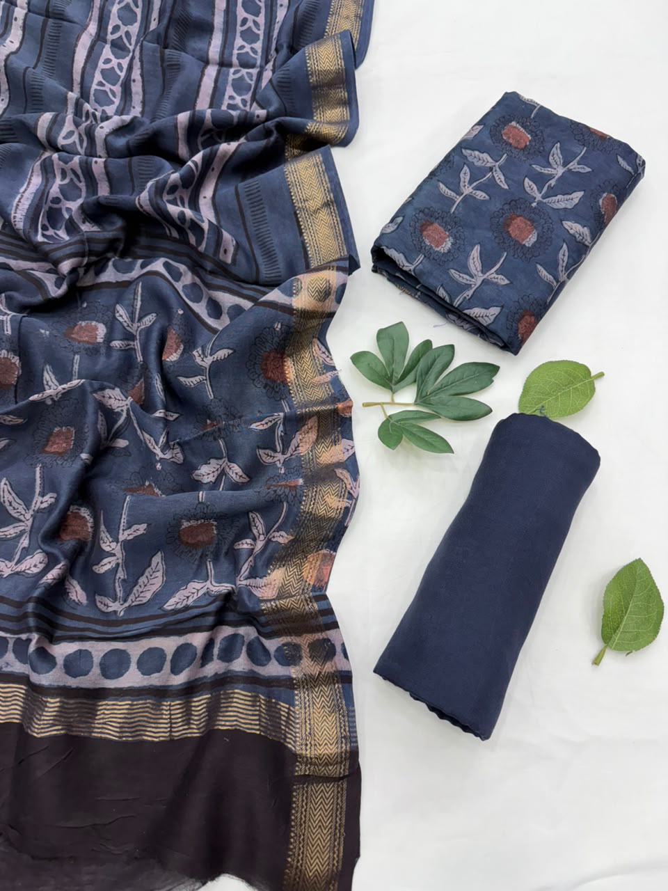 Folded and draped navy blue Maheshwari silk unstitched suit set with botanical block prints in beige, brown, and muted orange, styled on a white background with decorative green leaves