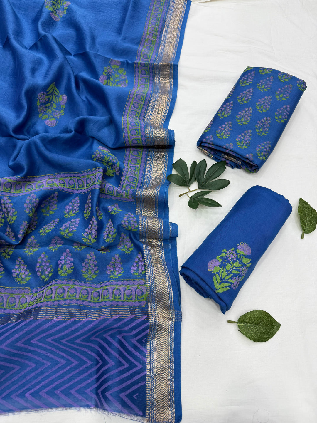 Blue patterned fabric set with a wide border and matching folded pieces, arranged on a white surface with green leaves as styling props.