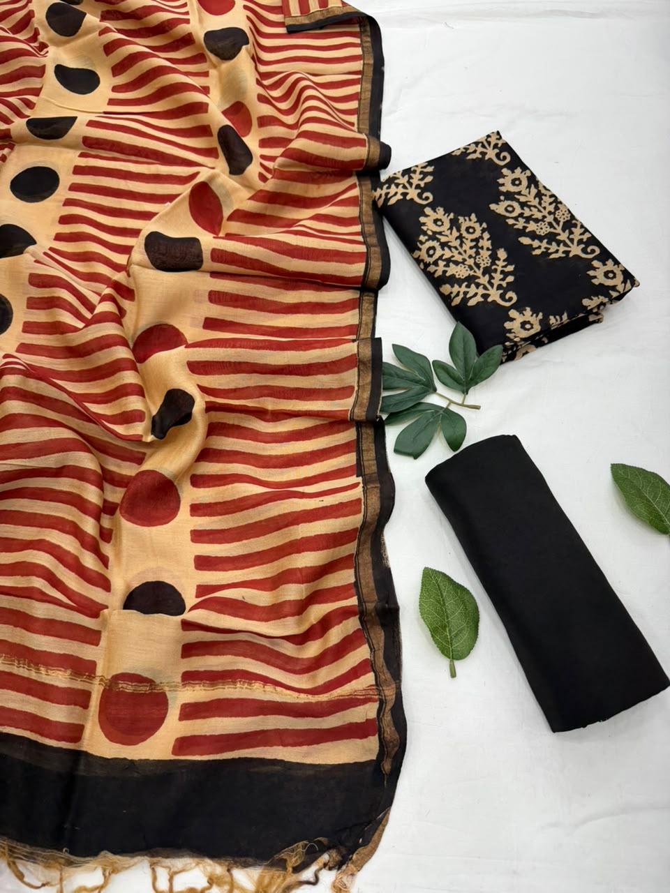 Flat lay view of a Bagru Syahi Hand Block Printed Chanderi Silk Suit Set featuring a folded black Chanderi silk fabric with gold floral block print, a matching black bottom fabric, and a cream dupatta with red and black block-printed motifs and a striped border