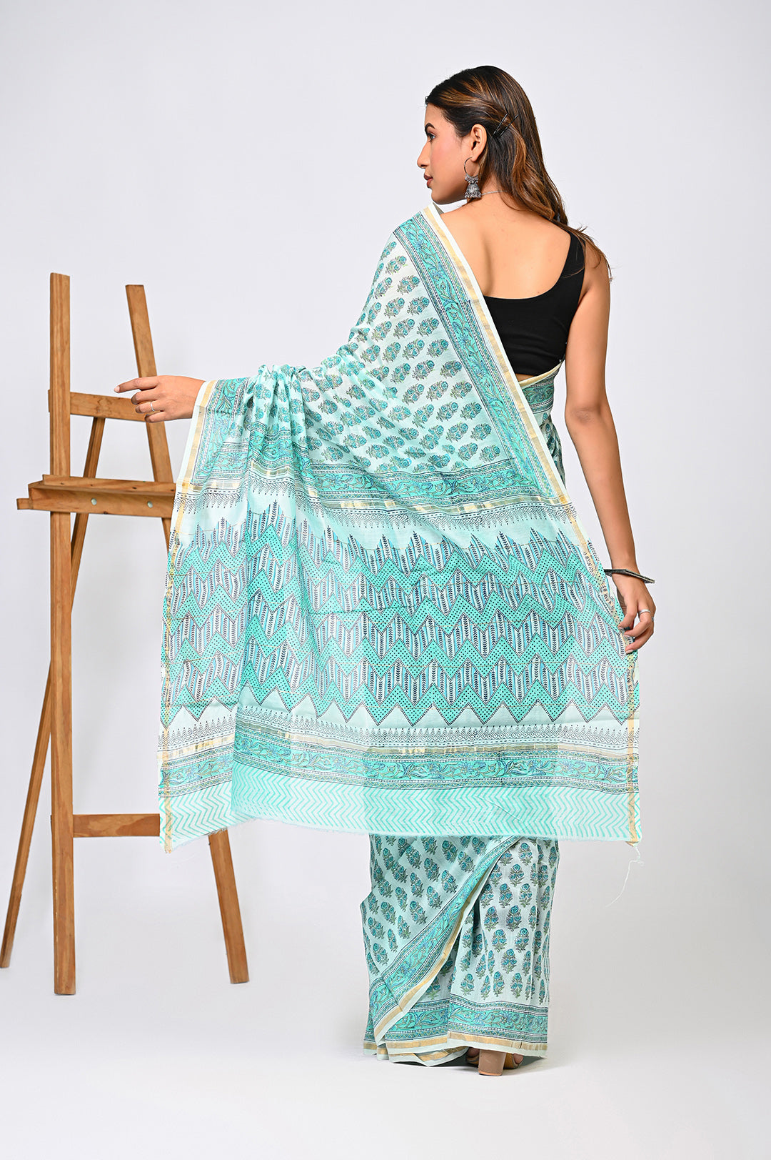 Back view of a woman modeling a light green Sanganeri print Chanderi Silk saree, showcasing its shimmering sheer drape, delicate hand block printed buti motifs, and matching patterned pallu next to a wooden easel.