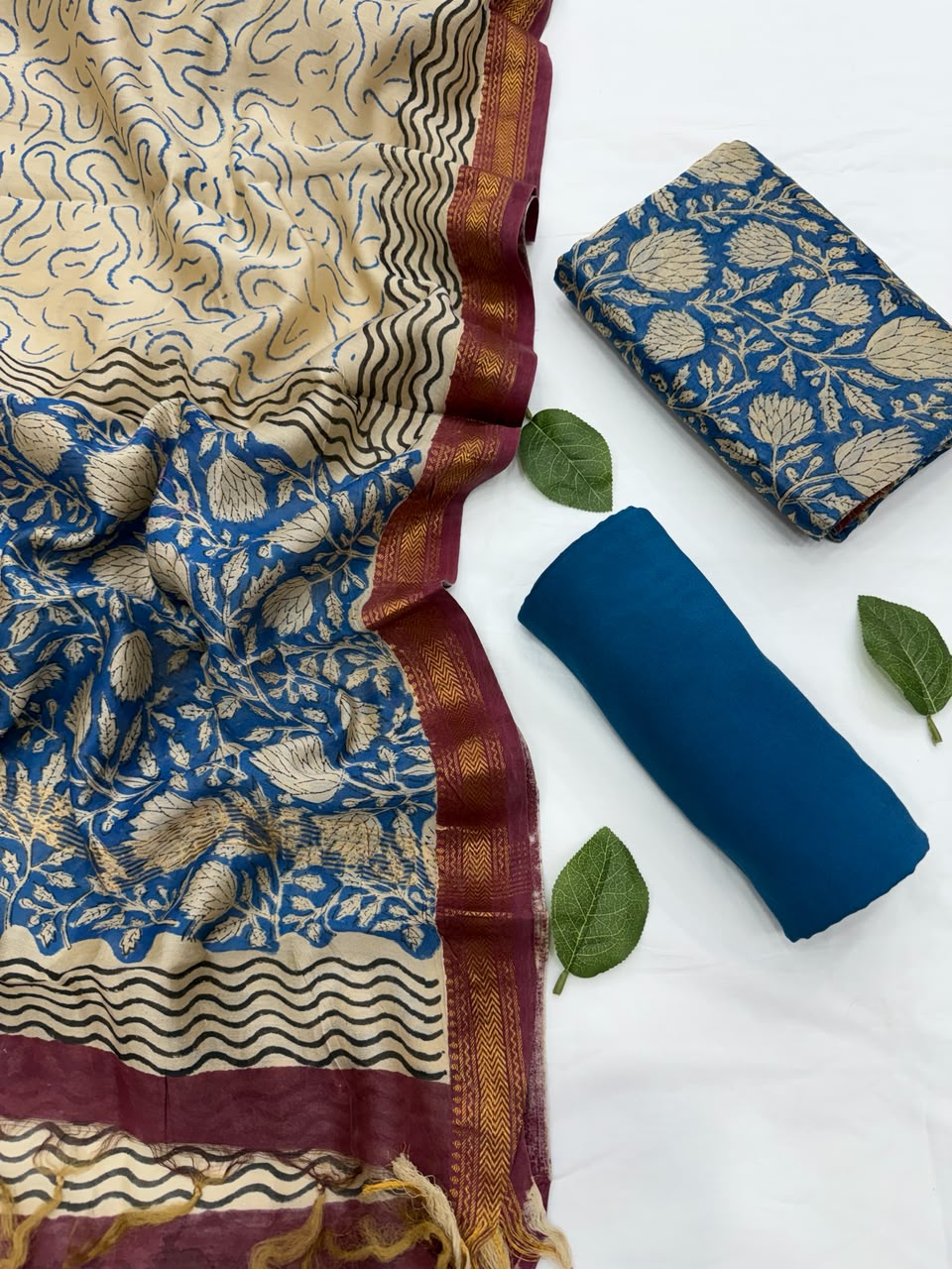 Full flat lay view of the Blue Bagru Hand Block Print Maheshwari Silk Salwar Suit Set, showing a beige and deep blue patterned dupatta with a rich maroon Zari border, a matching floral block-printed kurta fabric, and a neatly rolled solid blue bottom fabric styled with fresh green leaves.