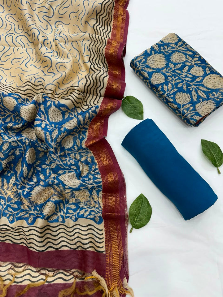 Full flat lay view of the Blue Bagru Hand Block Print Maheshwari Silk Salwar Suit Set, showing a beige and deep blue patterned dupatta with a rich maroon Zari border, a matching floral block-printed kurta fabric, and a neatly rolled solid blue bottom fabric styled with fresh green leaves.
