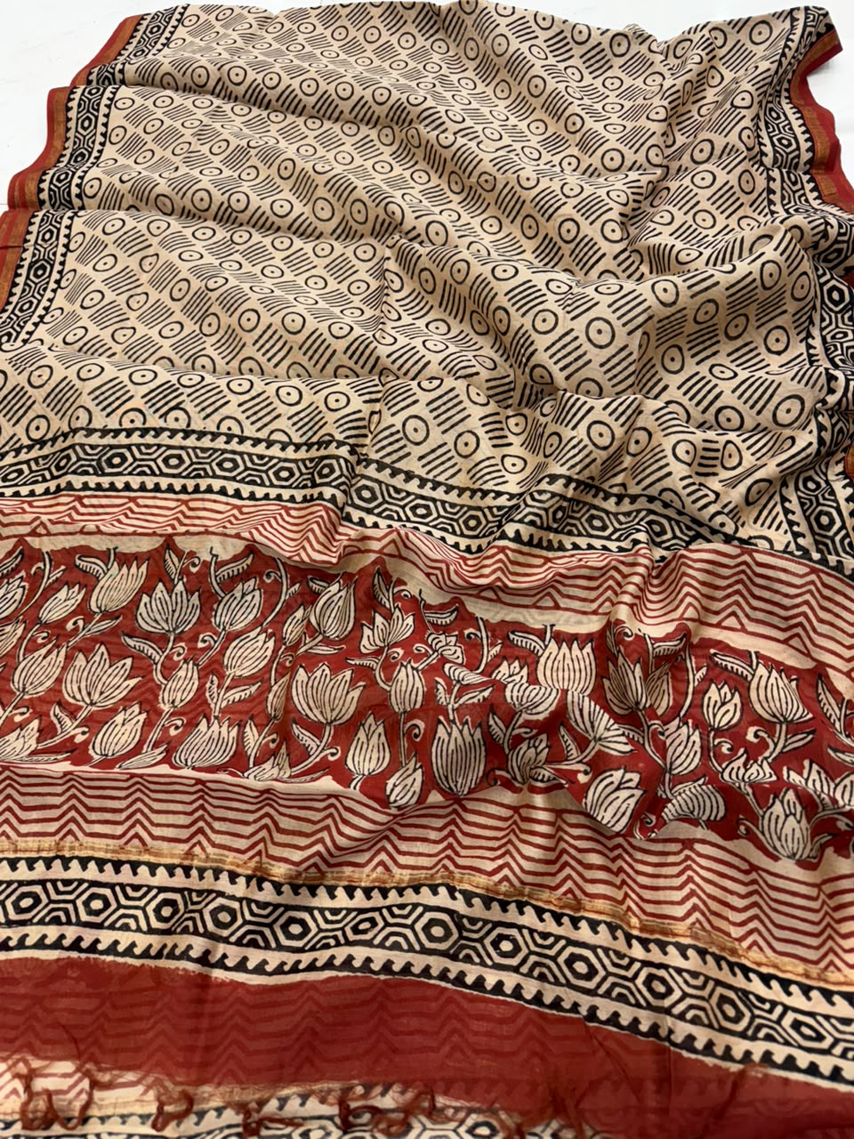 A close-up of a patterned textile, likely a sari or scarf, featuring a beige base with black geometric and floral block prints, and a wide border in rust red, beige, and black.