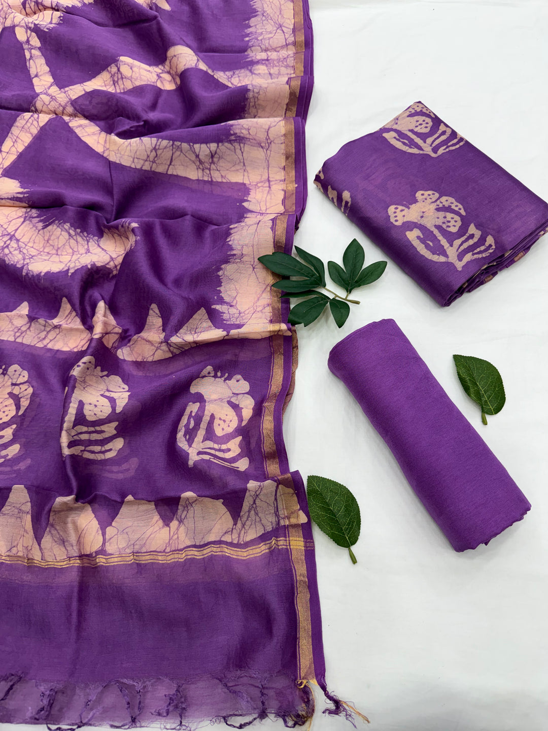 Purple cotton saree fabric with beige block-printed motifs and a gold border, shown alongside rolled and folded matching fabric pieces on a white background with green leaves.