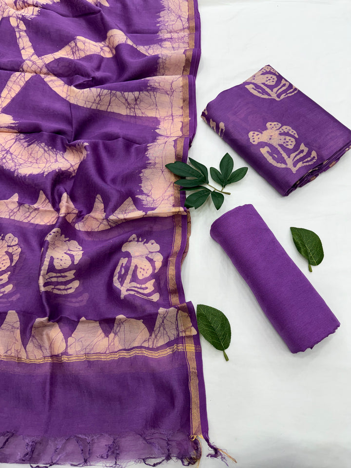 Purple cotton saree fabric with beige block-printed motifs and a gold border, shown alongside rolled and folded matching fabric pieces on a white background with green leaves.