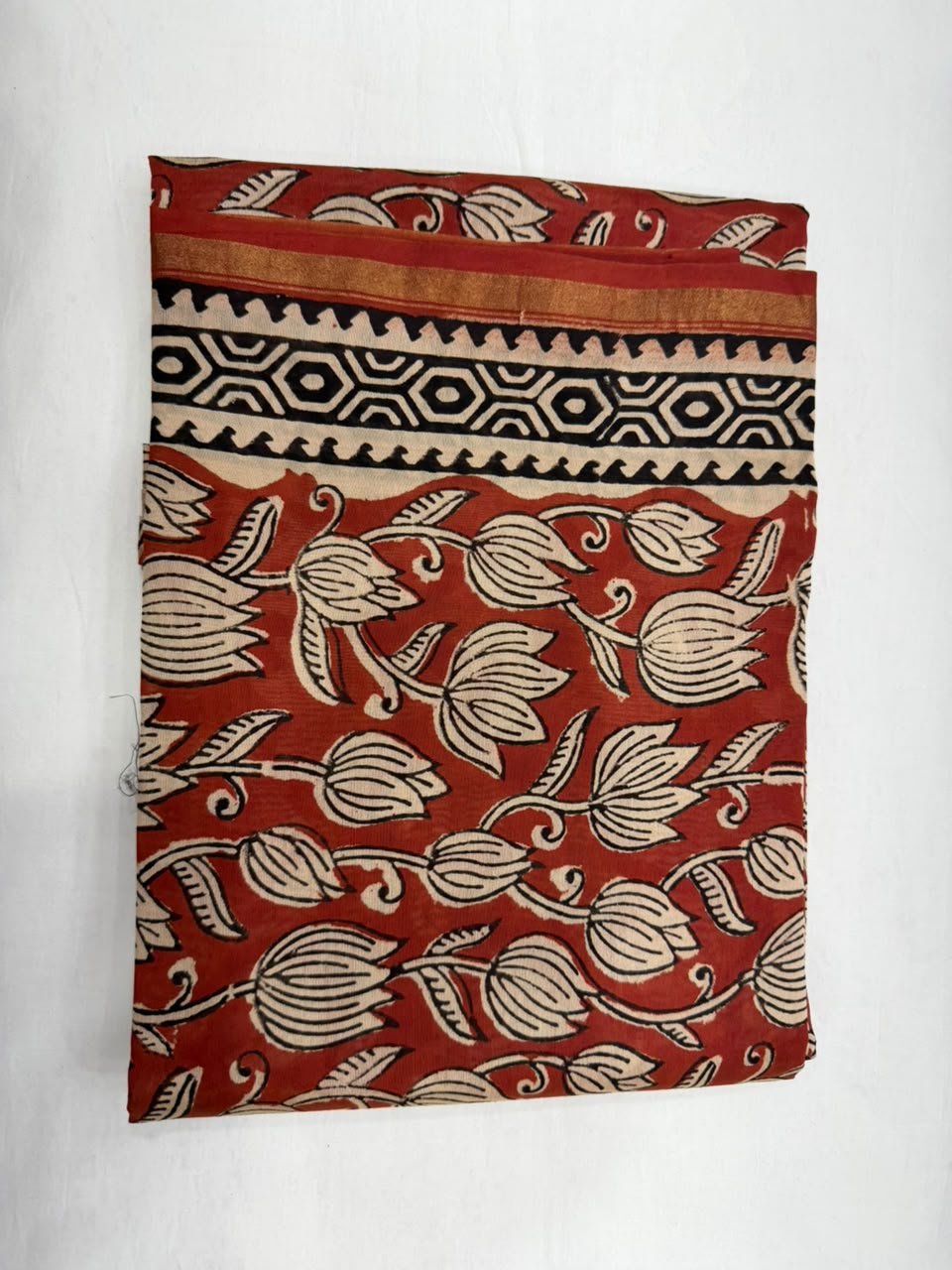 A folded piece of rust-red cotton fabric with a beige floral block print and a black geometric border pattern, displayed against a white background.
