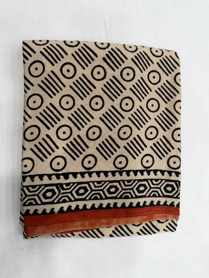 A folded textile, likely a saree, featuring a beige base with a black geometric pattern of circles, dots, and stripes, finished with a rust orange border.