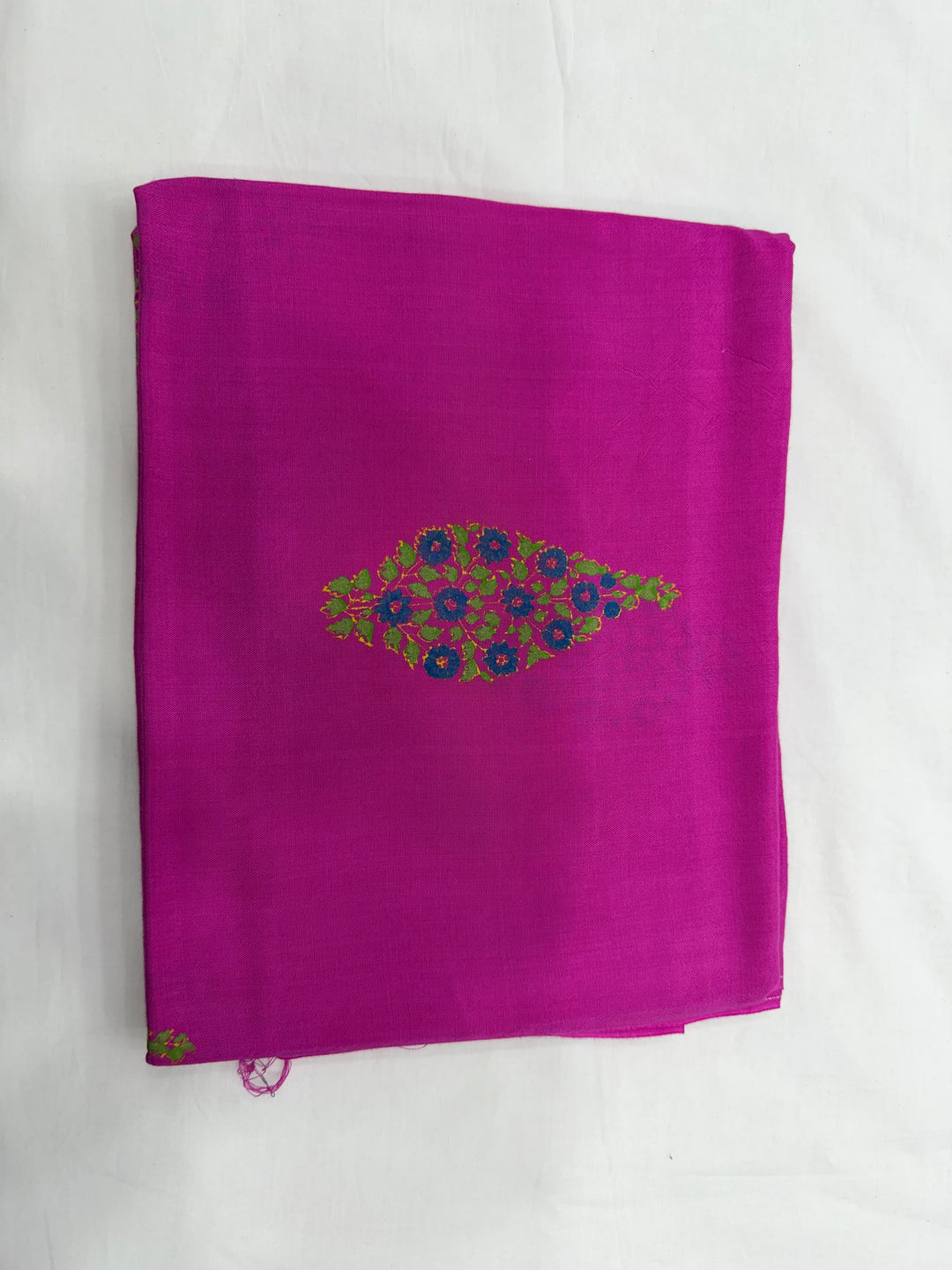 A folded magenta pink fabric, likely a saree or scarf, featuring a central embroidered peacock motif in green, blue, and purple on a white background.