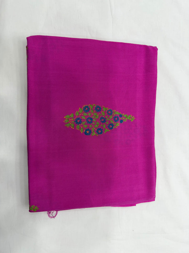 A folded magenta pink fabric, likely a saree or scarf, featuring a central embroidered peacock motif in green, blue, and purple on a white background.