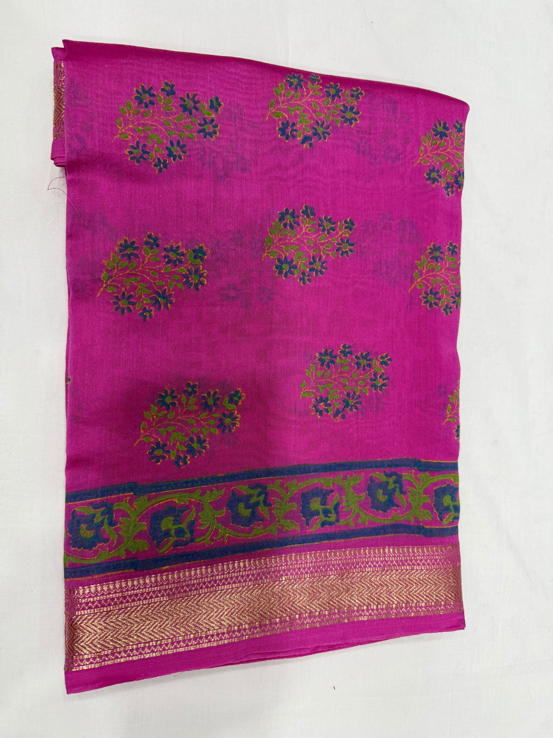 A folded magenta pink silk saree with woven green and blue floral motifs and a decorative border featuring gold zari detailing, laid flat on a white background.