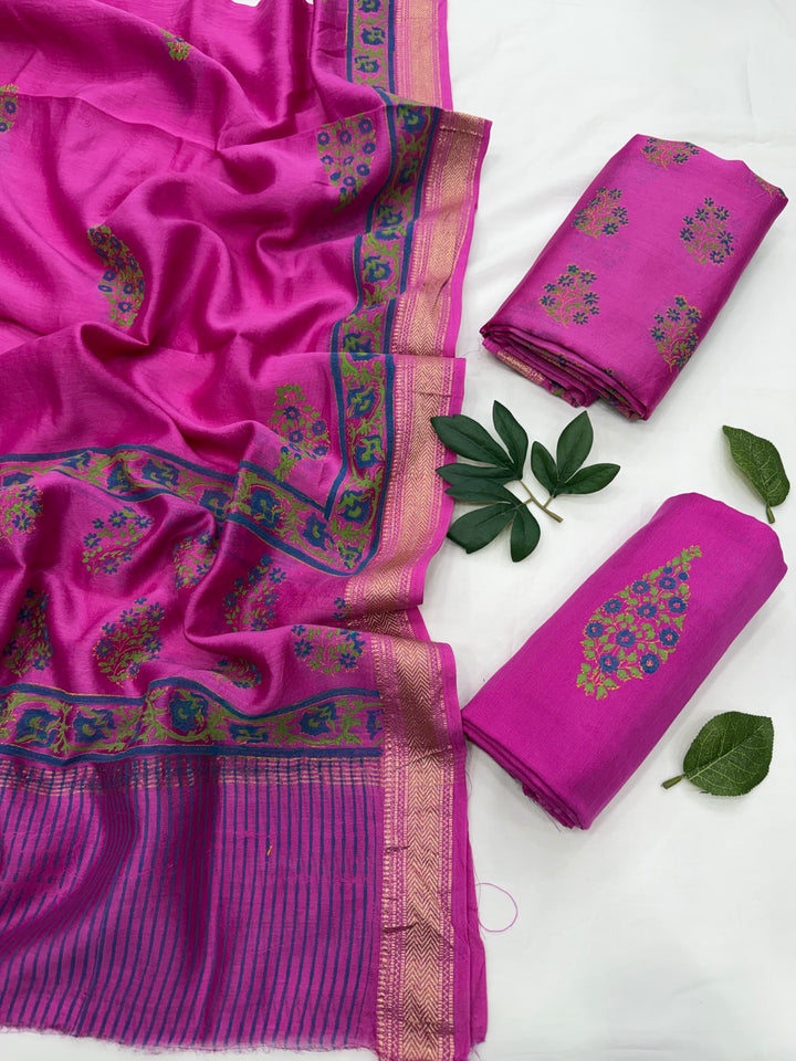 A folded and draped magenta pink Maheshwari silk suit set featuring a striped border and intricate floral block print motifs, displayed on a white background with decorative green leaves.