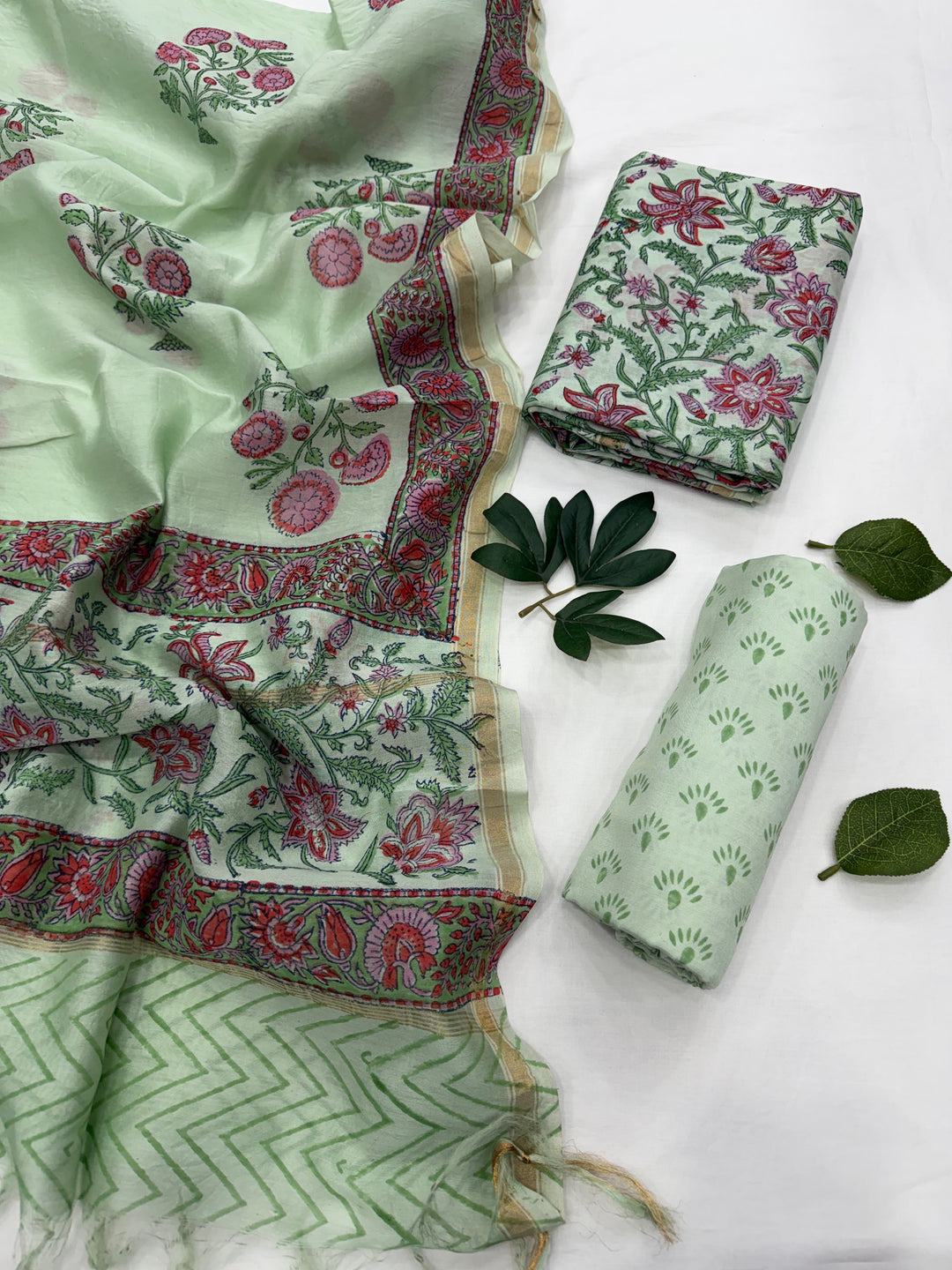 A folded and draped pastel green Chanderi silk suit set featuring delicate floral and geometric block prints in pink, red, and green, displayed against a clean white background.