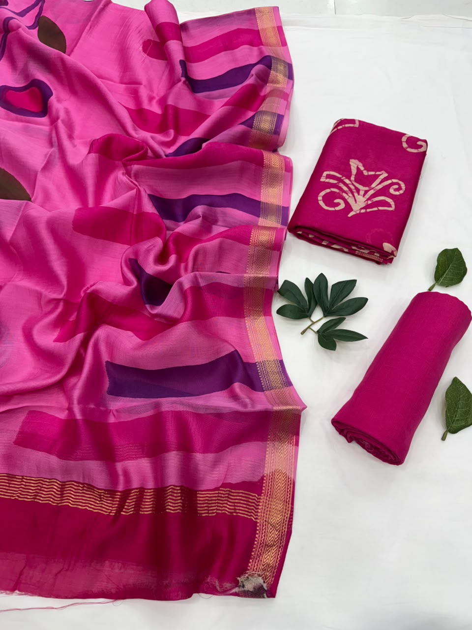 Pink batik print Maheshwari silk unstitched suit set featuring purple and magenta stripes with a gold border on the dupatta, displayed with a matching fabric piece and green decorative leaves on a white background
