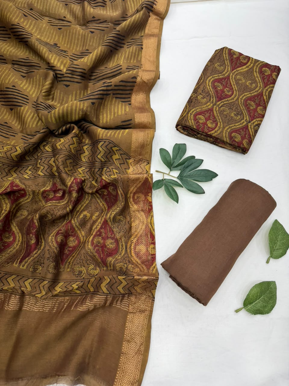 Brown and black checked Maheshwari silk unstitched suit set featuring a geometric pattern, rich gold zari border, and zigzag printed accents, displayed alongside folded matching fabric pieces and green leaves on a white background