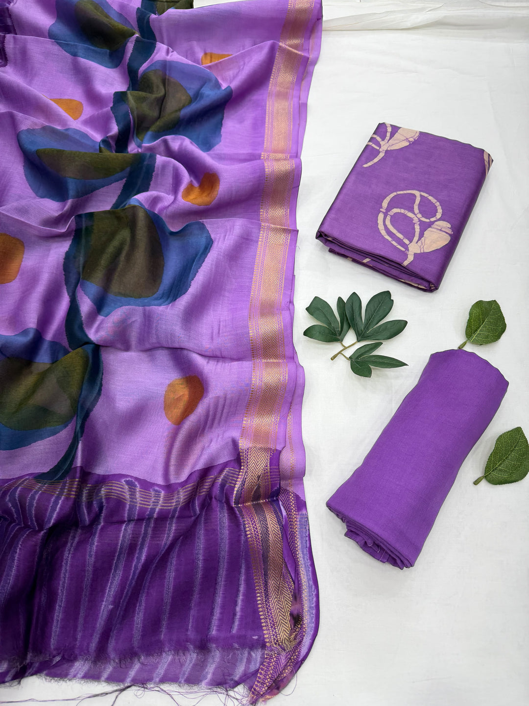 Purple designer handblock print Maheshwari silk unstitched suit set featuring abstract floral prints and a rich gold zari border, displayed alongside folded matching fabric pieces and decorative green leaves on a white background