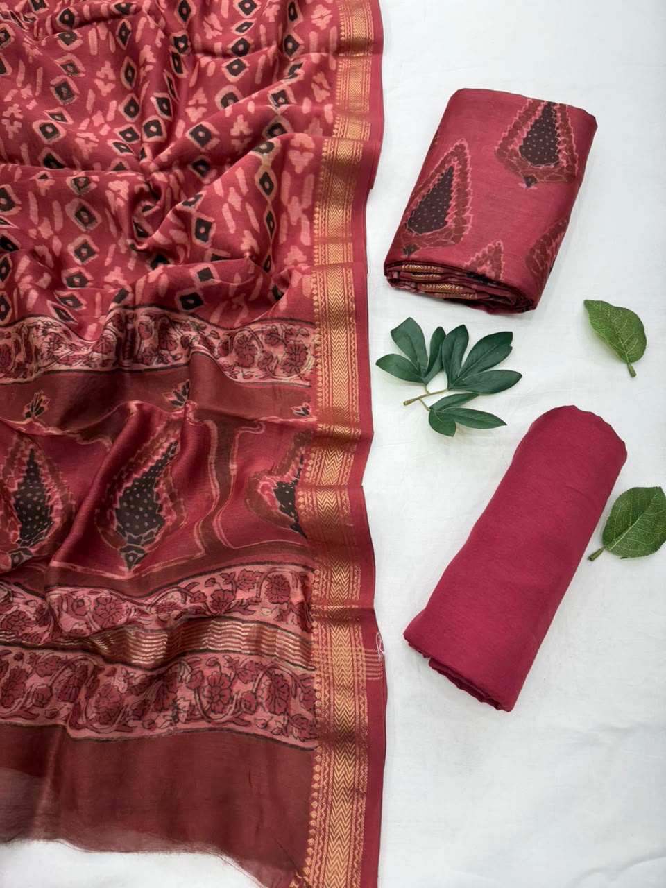 A folded and draped maroon Maheshwari silk suit set featuring intricate Vanspati block print motifs, a contrasting red woven zari border, and green leaf accents displayed on a white background.