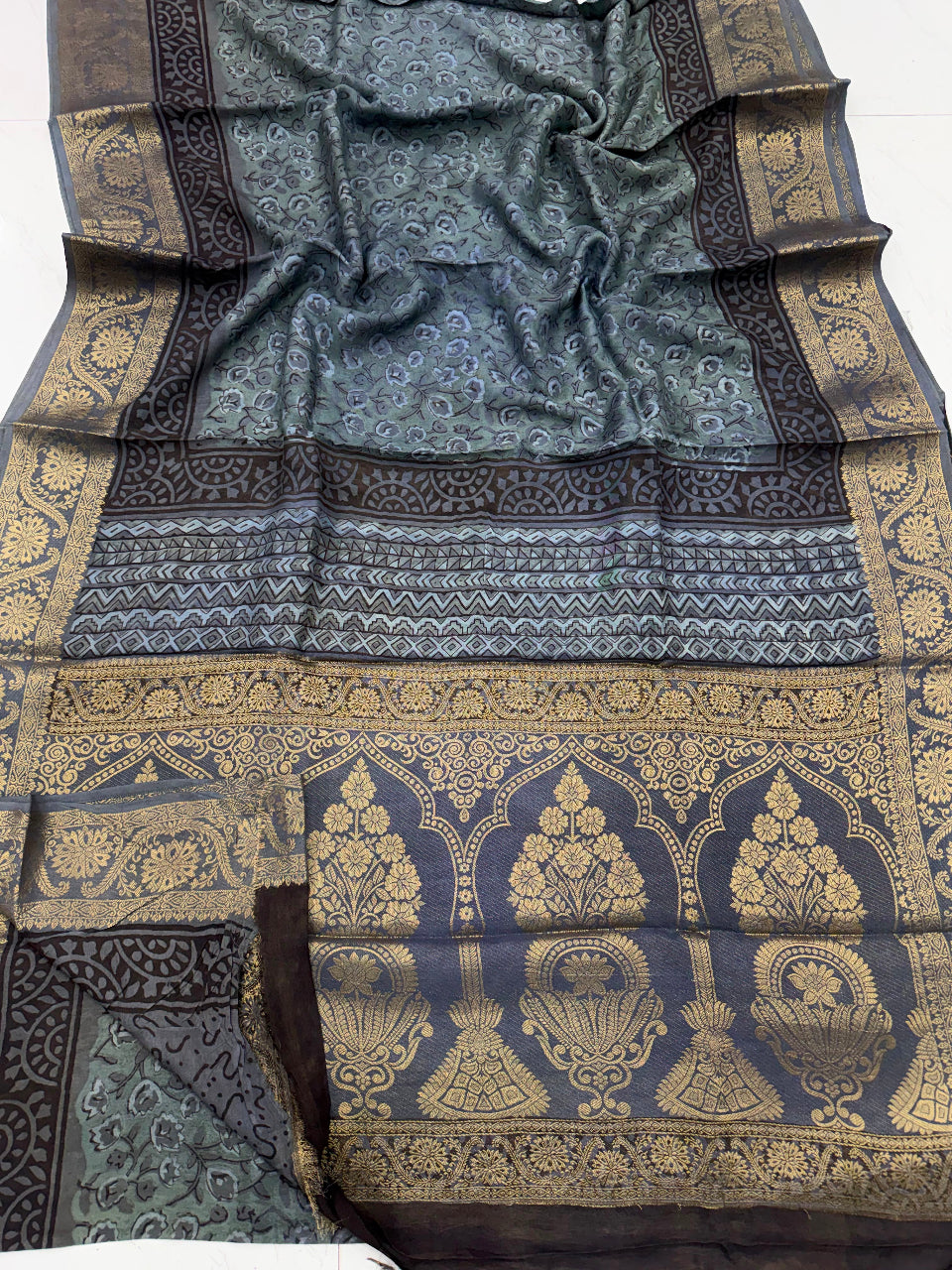 A dark slate blue and black silk saree with intricate gold zari embroidery, featuring a wide decorative border with arch motifs and a patterned body.