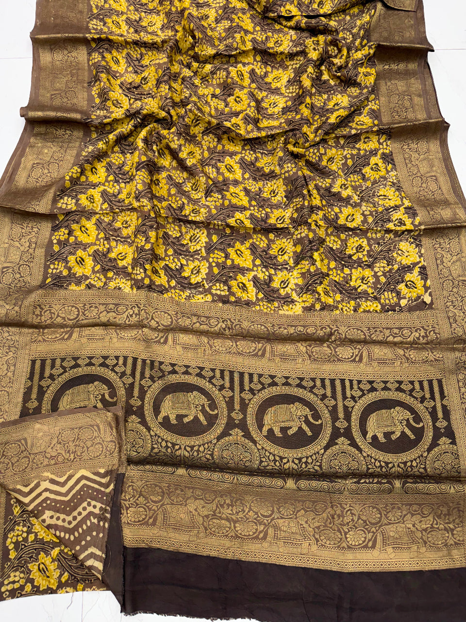 A dark brown and yellow printed fabric, likely a saree, featuring a floral pattern on the body and a wide decorative border with elephant motifs.