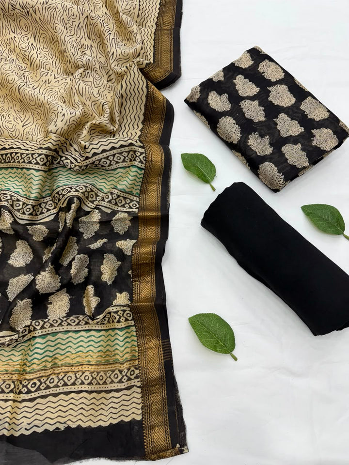 Full flat lay view of the Bagru Buti Print Natural Black Maheshwari Silk Suit Set, showcasing the black kurta fabric with golden floral motifs, a matching patterned dupatta with a golden woven Zari border, and a neatly rolled solid black bottom fabric decorated with fresh green leaves.