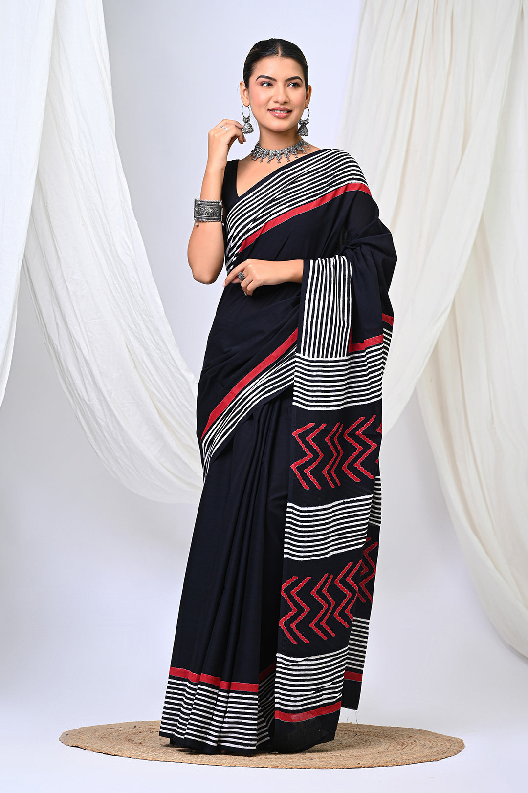 Full-length front view of a model in a premium plain black Bagru handblock print mulmul cotton saree, featuring a crisp black and white stripe border and natural red geometric motifs.