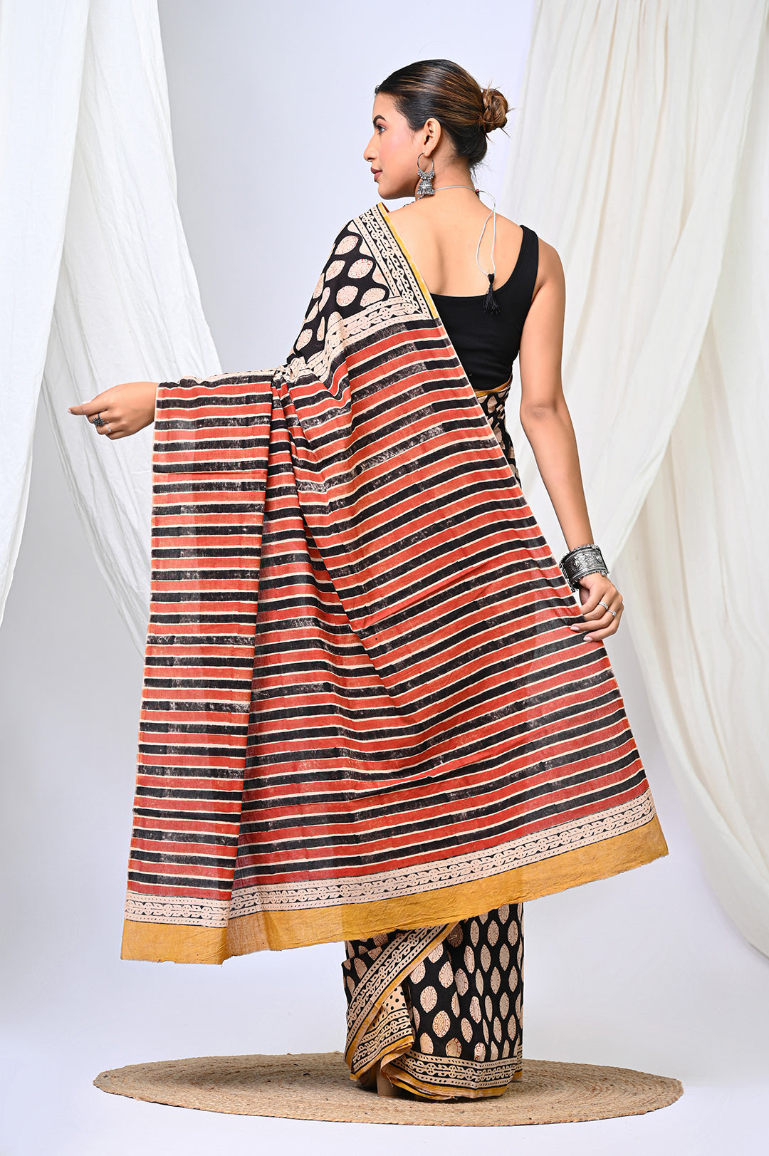 Full-length rear view of a model showcasing the elegant drape of a black Bagru handblock print mulmul cotton saree, featuring a long pallu with rust, black, and cream horizontal stripes and a mustard-gold hem border.