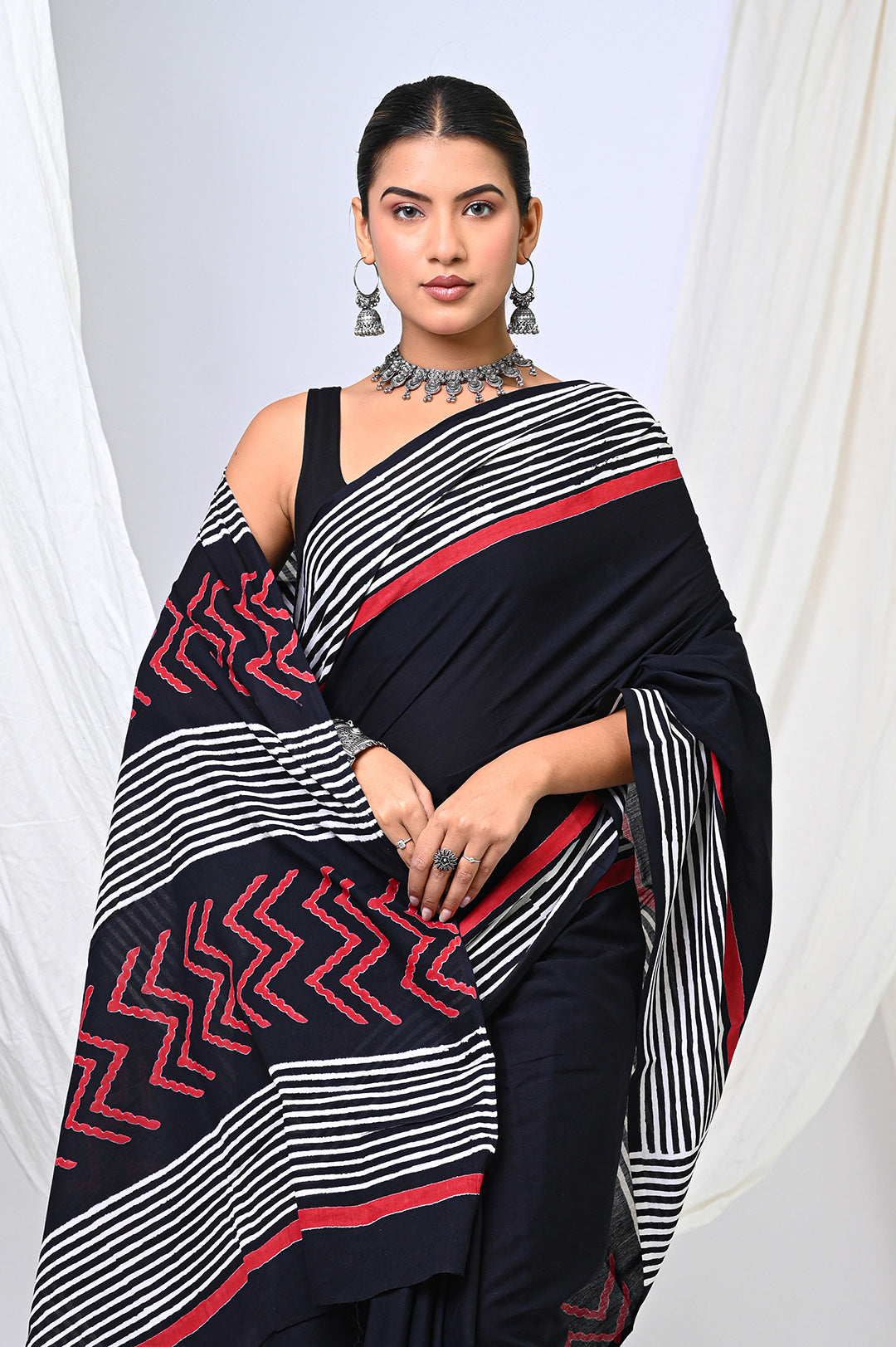 Close-up studio shot of a woman draping a traditional natural black dye Rajasthani block print cotton saree with a graphic stripe border, styled with an oxidized silver statement necklace against a white backdrop.