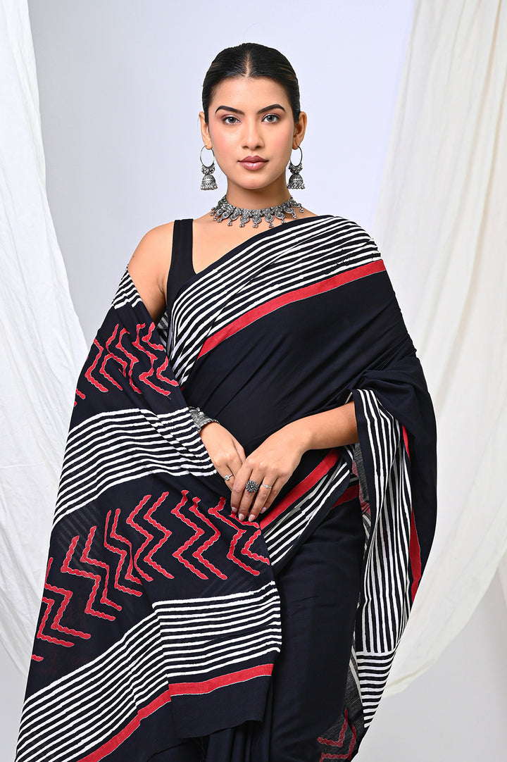 Close-up studio shot of a woman draping a traditional natural black dye Rajasthani block print cotton saree with a graphic stripe border, styled with an oxidized silver statement necklace against a white backdrop.