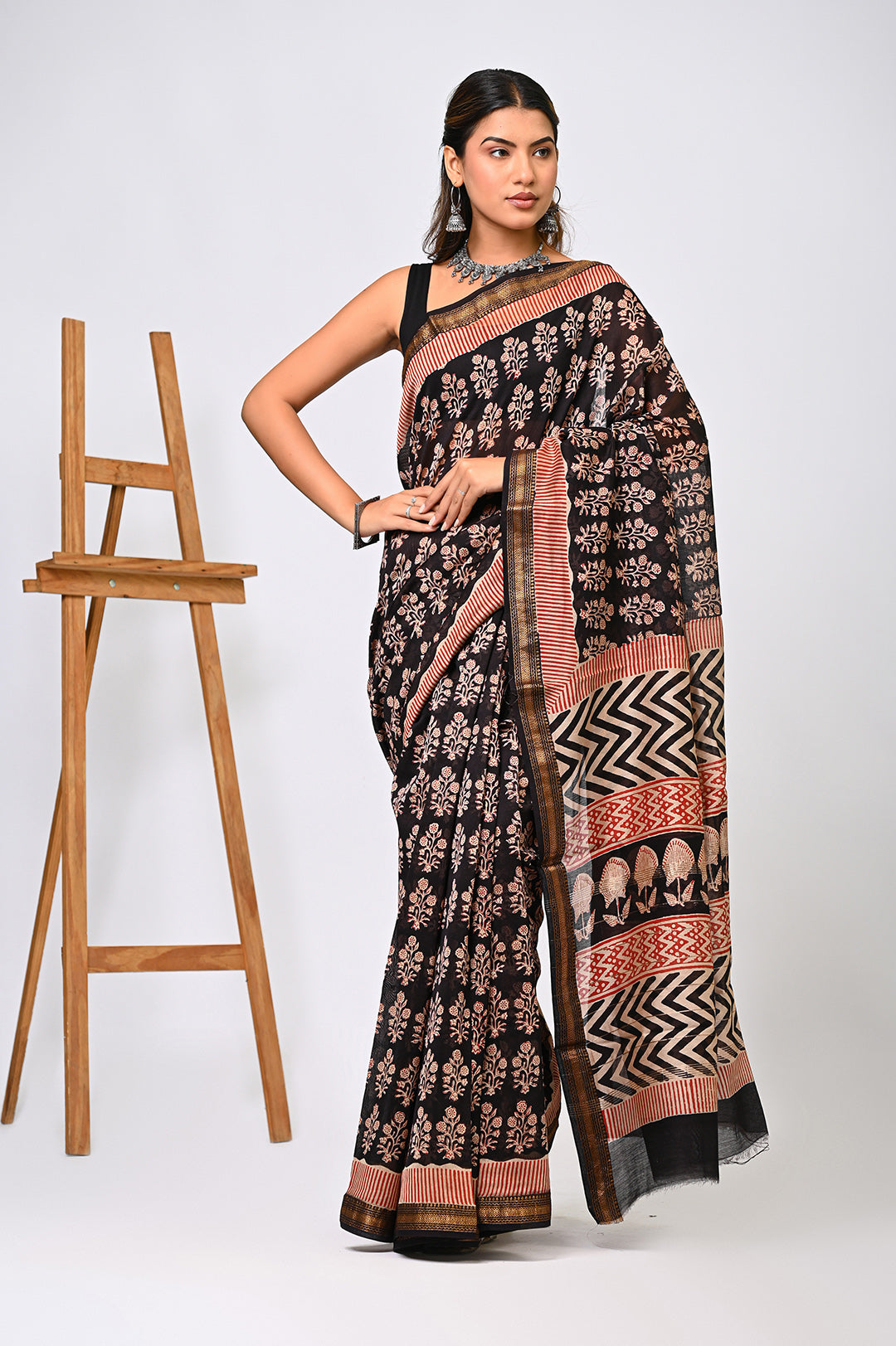 A woman modeling a natural black Maheshwari silk saree with an all-over floral buti pattern in rust and cream, featuring a striking black and white zigzag border.