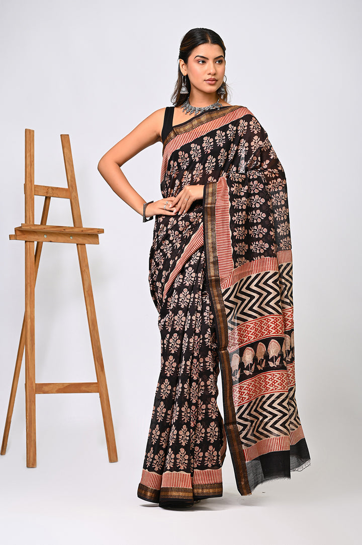 A woman modeling a natural black Maheshwari silk saree with an all-over floral buti pattern in rust and cream, featuring a striking black and white zigzag border.