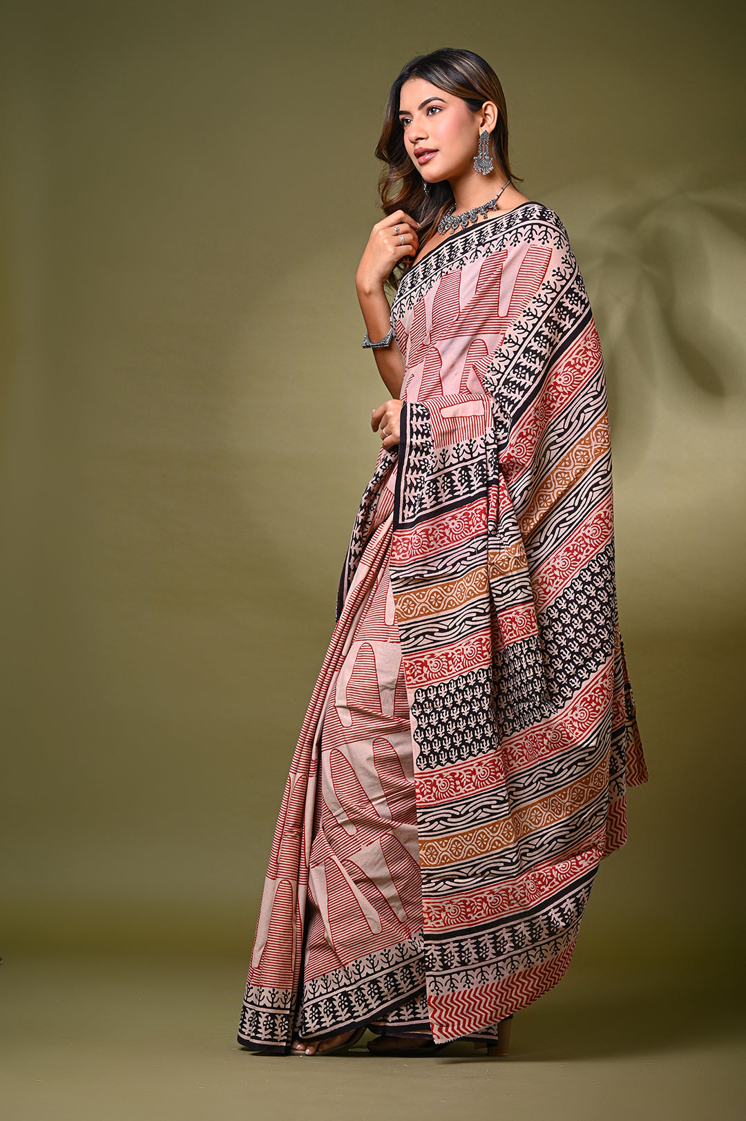 Studio shot of a woman draping a traditional Rajasthani block print cotton saree in natural red and off-white tones, paired with a black sleeveless blouse against an olive green background.