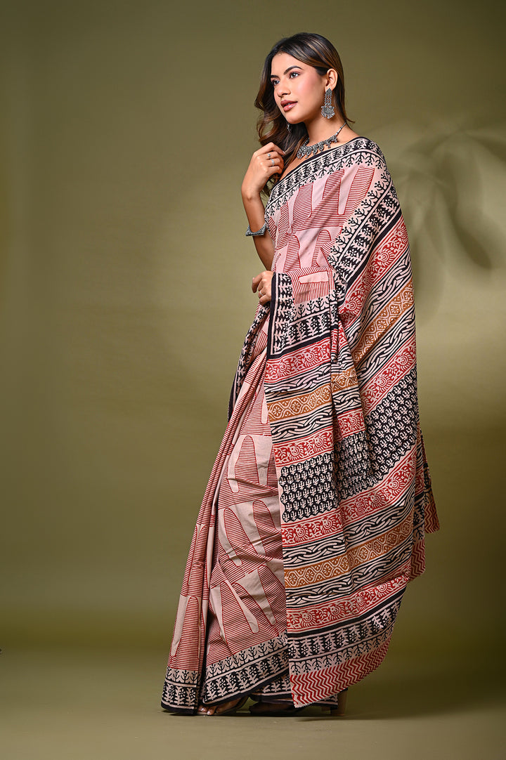Studio shot of a woman draping a traditional Rajasthani block print cotton saree in natural red and off-white tones, paired with a black sleeveless blouse against an olive green background.