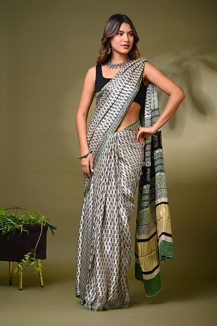 A woman modeling a natural cream Modal Silk saree with an all-over black and grey handblock printed buti motif, featuring a green salvage edge and a traditional patterned pallu against an olive green backdrop.