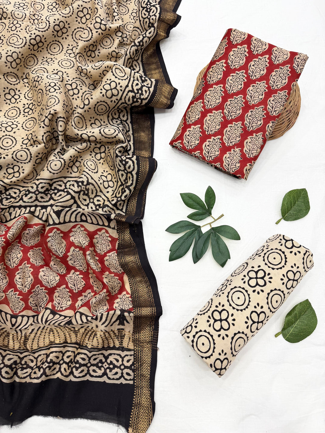 A folded and draped textile set with a beige and black floral block print, a red and beige patterned fabric piece, and a matching rolled beige fabric piece with black motifs, styled with green leaves on a white background.