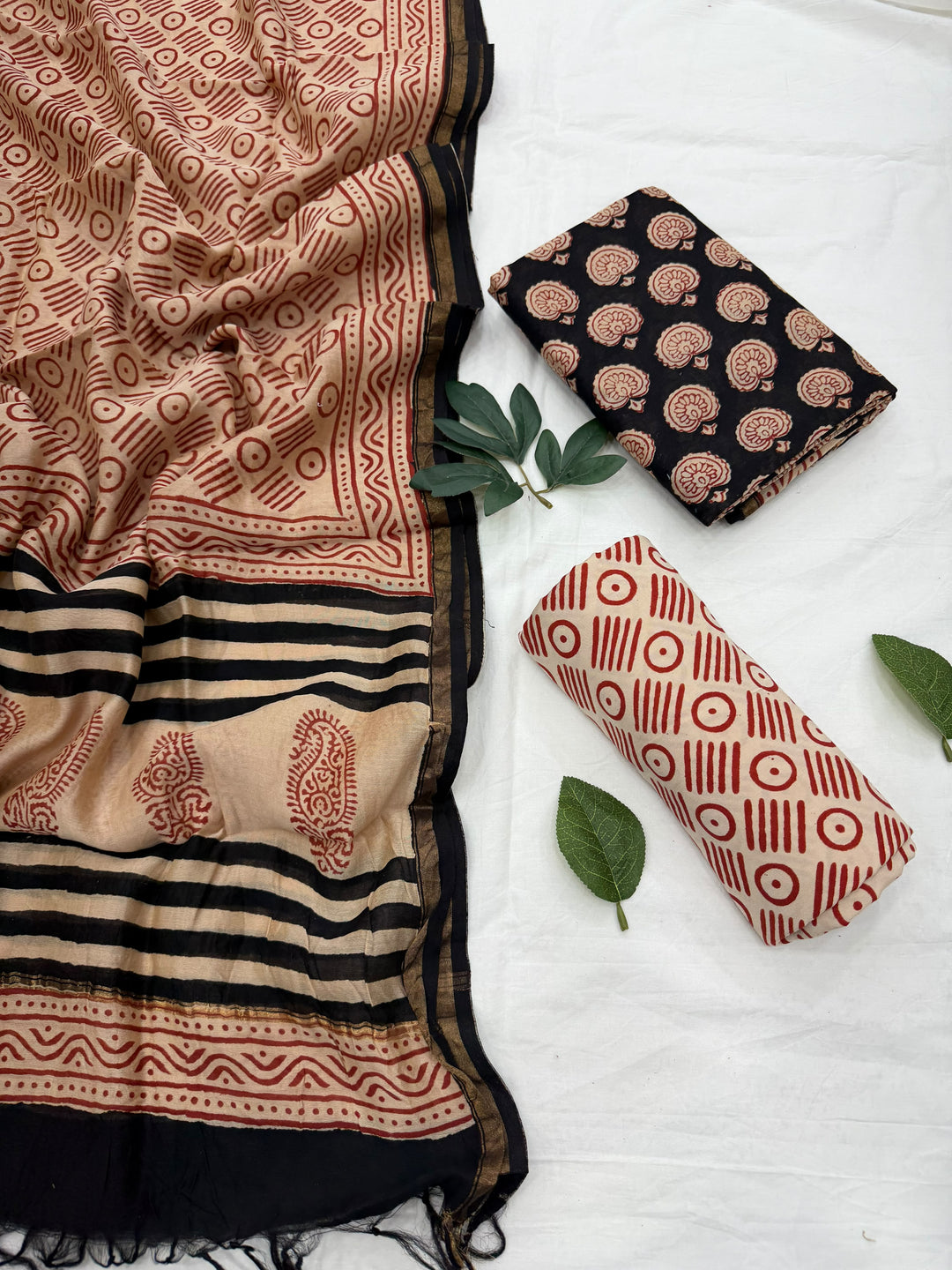 A folded and draped fabric set with three distinct prints: a large beige and black striped patterned piece, a folded black piece with beige paisley motifs, and a rolled cream piece with red geometric patterns. Styled with green leaves on a white fabric background.