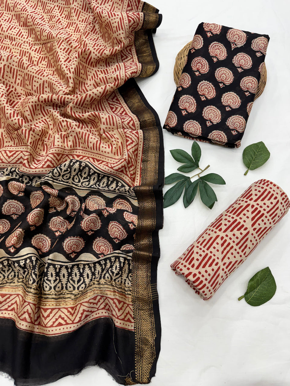 A folded and rolled textile set with a red and beige block-print pattern and a black border, displayed on a white background with decorative green leaves. The set includes a draped fabric piece, a folded square piece, and a rolled cylindrical piece.