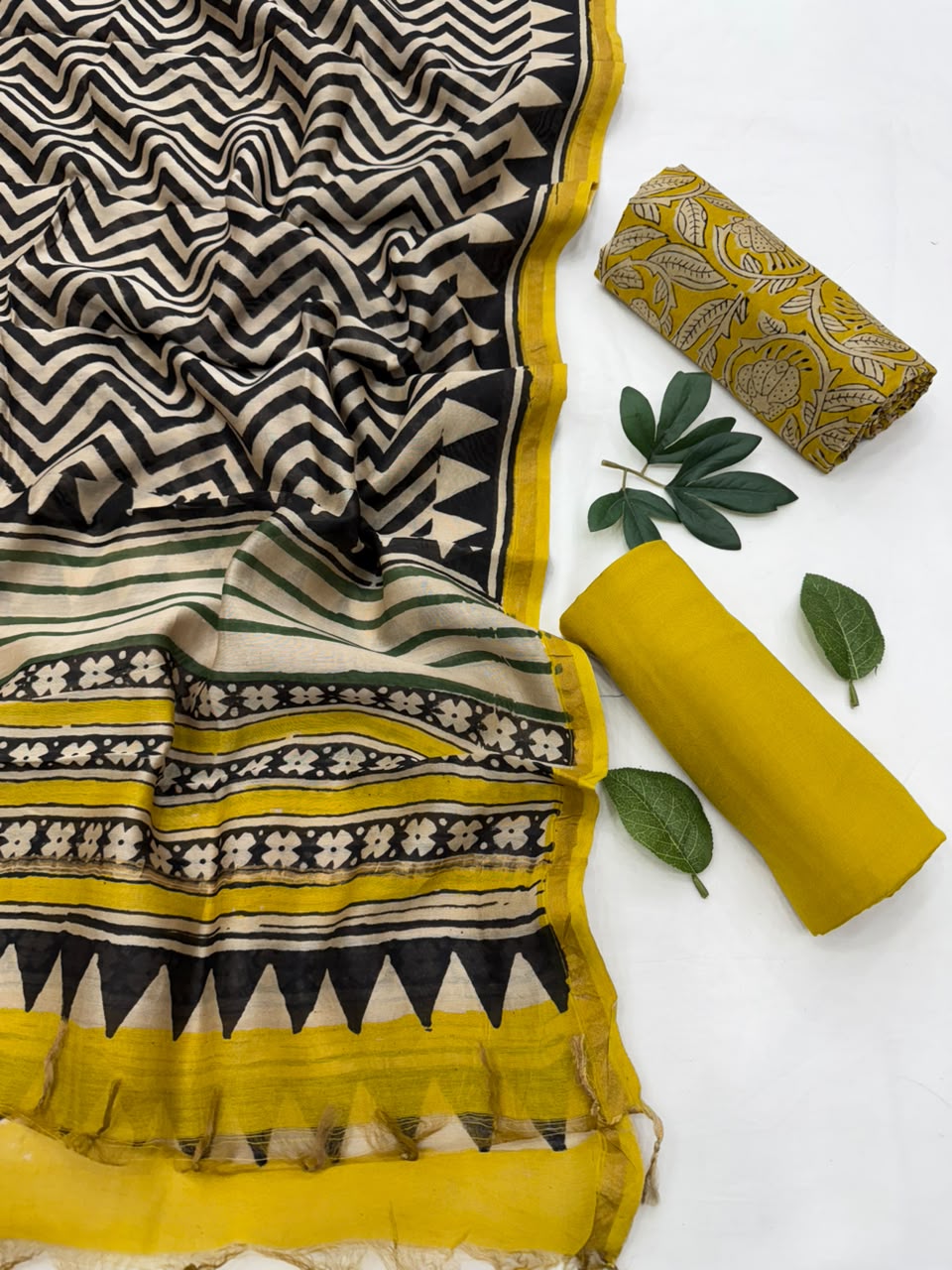 A folded and rolled mustard yellow and black printed fabric set, likely a saree or scarf, displayed on a white background with decorative green leaves. The fabric features a black and beige chevron pattern on one section, and a border with stripes, flowers, and triangles in yellow, black, and beige.