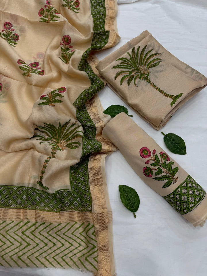 Full flat lay view of the Beige Maheshwari Silk Suit Set, featuring a kurta fabric with pink floral prints and a dark green border, a matching dupatta with intricate palm tree and chevron patterns, and a rolled beige bottom fabric styled with fresh green leaves.