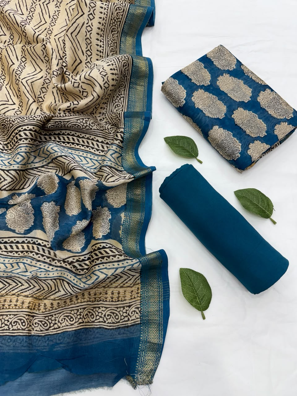 Full flat lay view of the Blue Traditional Bagru Hand Block Print Maheshwari Silk Suit Set, displaying a folded beige and blue patterned dupatta with a golden Zari border, a matching blue hand block-printed kurta fabric, and a neatly rolled solid deep blue bottom fabric styled with fresh green leaves.
