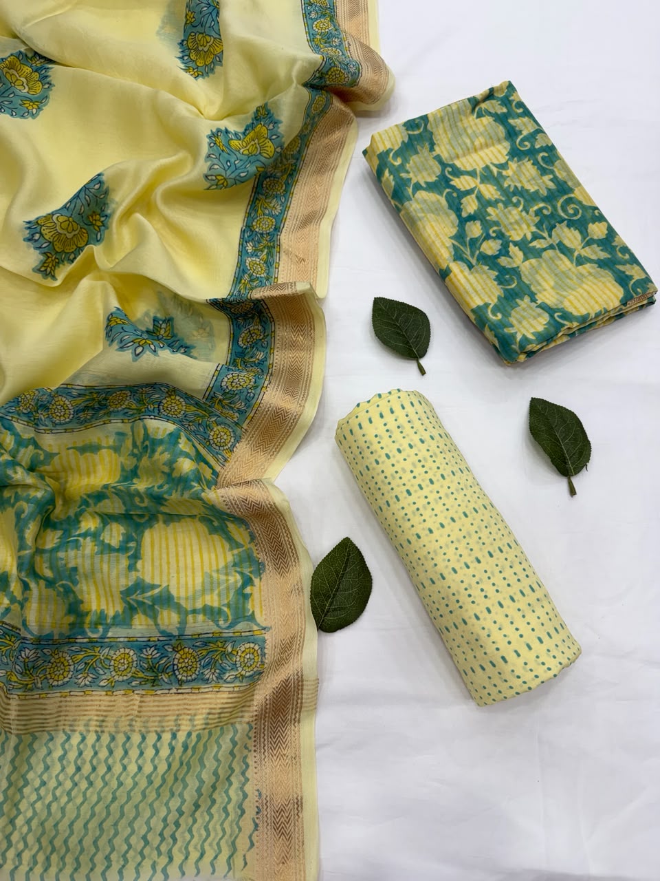 Full flat lay view of the Bright Yellow Hand Block Print Maheshwari Silk Suit Set, showcasing the yellow polka-dotted kurta fabric, a matching translucent dupatta with green floral motifs and a golden Zari border, and a neatly folded green patterned bottom fabric styled with fresh leaves.