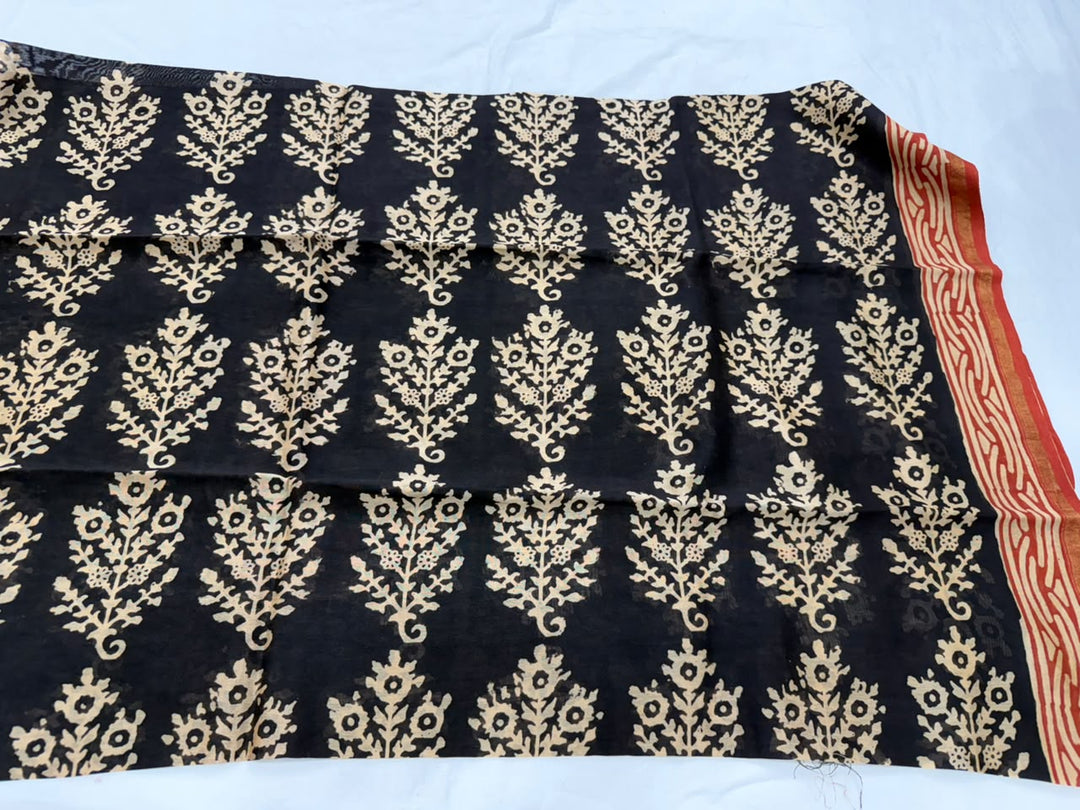 A black fabric piece, likely a saree or dupatta, featuring a repeating beige floral and leaf block print and a red and beige patterned border at the bottom, laid flat against a white background.