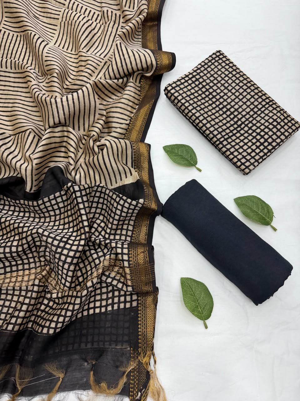 Full flat lay view of the Cream Bagru Checks Block Print Maheshwari Silk Suit Set, displaying a cream and black checked kurta fabric, a matching patterned dupatta with an elegant golden handloom Zari border, and a neatly rolled solid black bottom fabric styled with fresh green leaves.