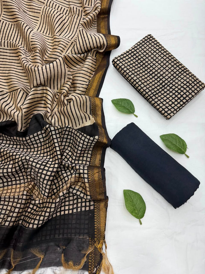 Full flat lay view of the Cream Bagru Checks Block Print Maheshwari Silk Suit Set, displaying a cream and black checked kurta fabric, a matching patterned dupatta with an elegant golden handloom Zari border, and a neatly rolled solid black bottom fabric styled with fresh green leaves.