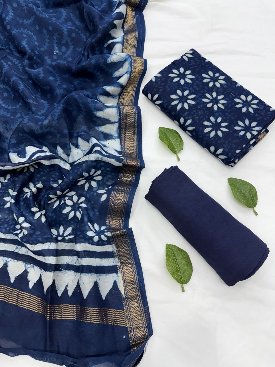 Full flat lay view of the Dabu Print Indigo Maheshwari Silk Suit Set, showcasing the indigo blue hand block-printed kurta fabric, matching patterned dupatta with a golden woven Zari border, and solid deep blue bottom fabric decorated with fresh green leaves.