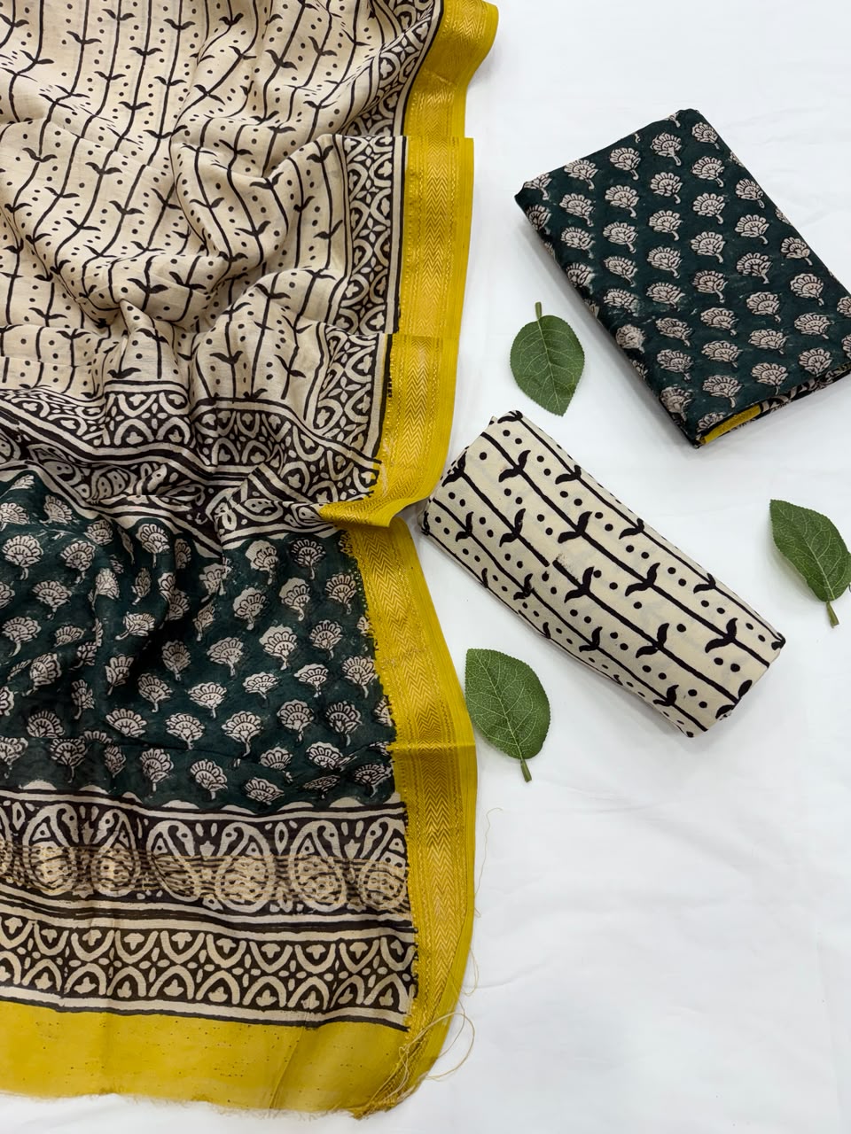 Full flat lay view of the Dark Green Bagru Buti Print Maheshwari Silk Salwar Suit Set, displaying a folded cream dupatta with black block prints and a golden yellow Zari border, alongside a dark green block-printed kurta fabric, a cream patterned bottom fabric, and fresh green leaves.