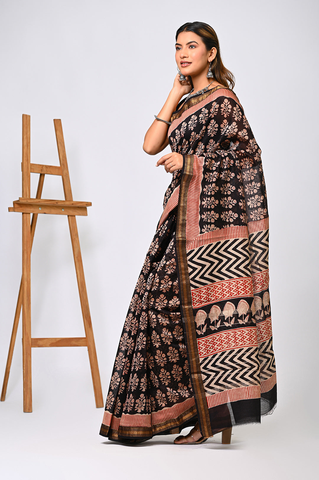An authentic hand block printed Maheshwari silk saree in organic black and earthy rust tones, styled elegantly next to a wooden artist easel backdrop.