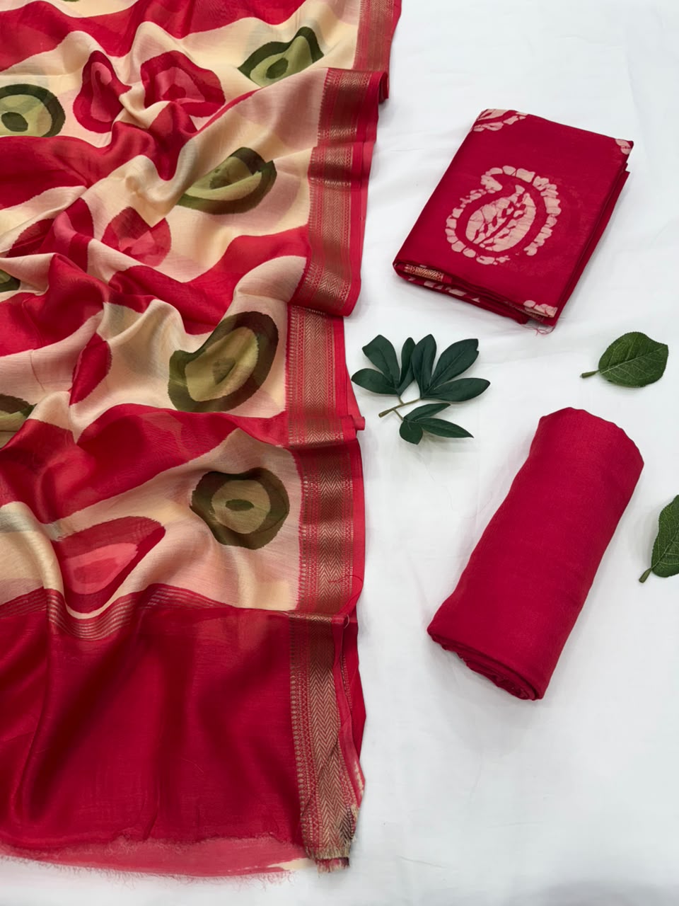 A folded and draped red saree or fabric set displayed on a white surface, with one section spread out to show a cream base, red floral and leaf motifs, and a gold border, alongside folded red pieces and green leaves as styling props.